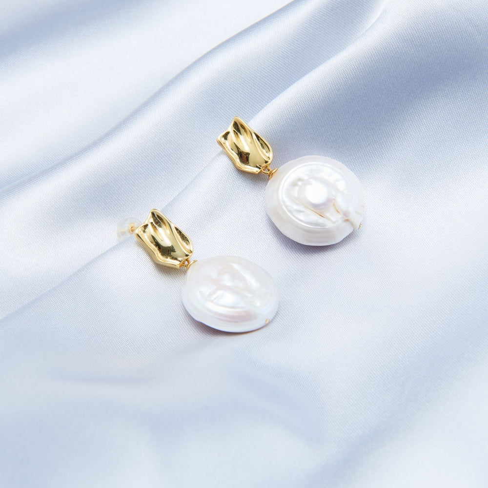 
                      
                        Elegant Pearlra modern baroque earrings with round pearls and gold accents on soft fabric
                      
                    