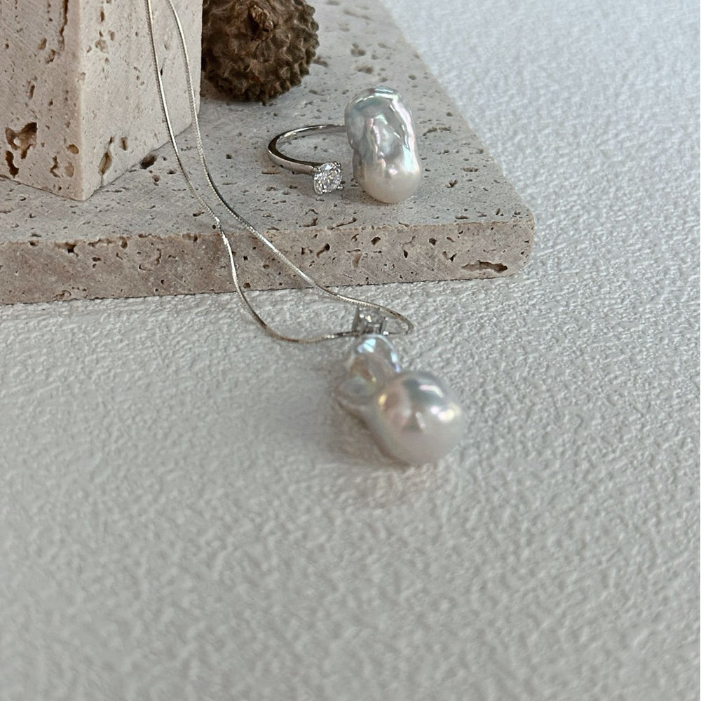 
                      
                        Petite baroque pearl ring showcased next to a silver necklace on a stone surface
                      
                    
